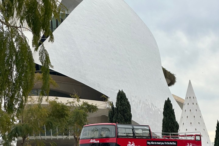 Red double-decker tour bus in front of modern white building with trees around.