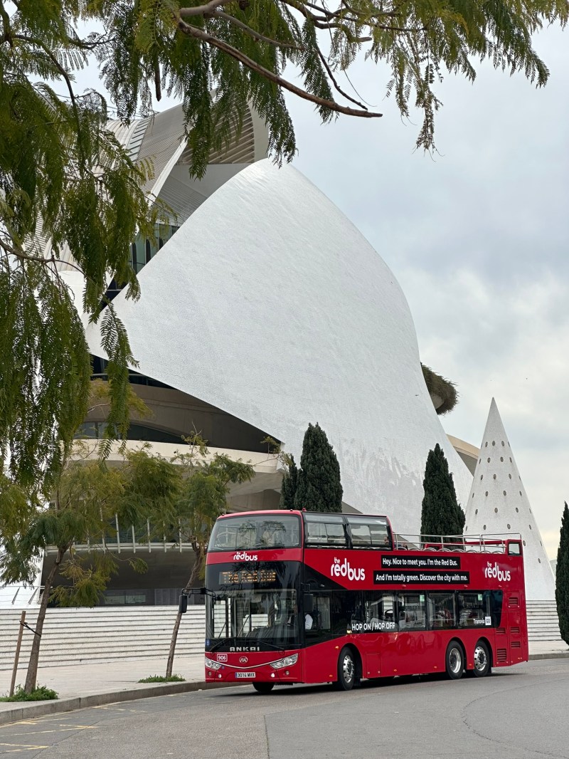 Red double-decker tour bus in front of modern white building with trees around.